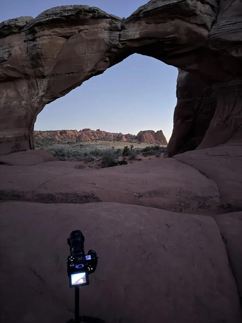 A camera on a low tripod on slickrock, screen glowing, aimed through the Broken Arch opening at deep blue twilight