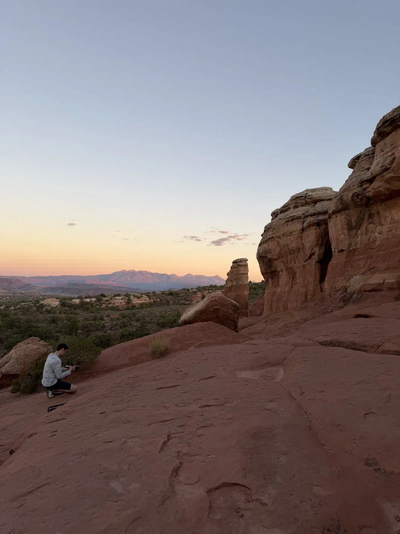 Nicholas crouched on slickrock with a camera, distant mountains catching pink sunset light on the horizon