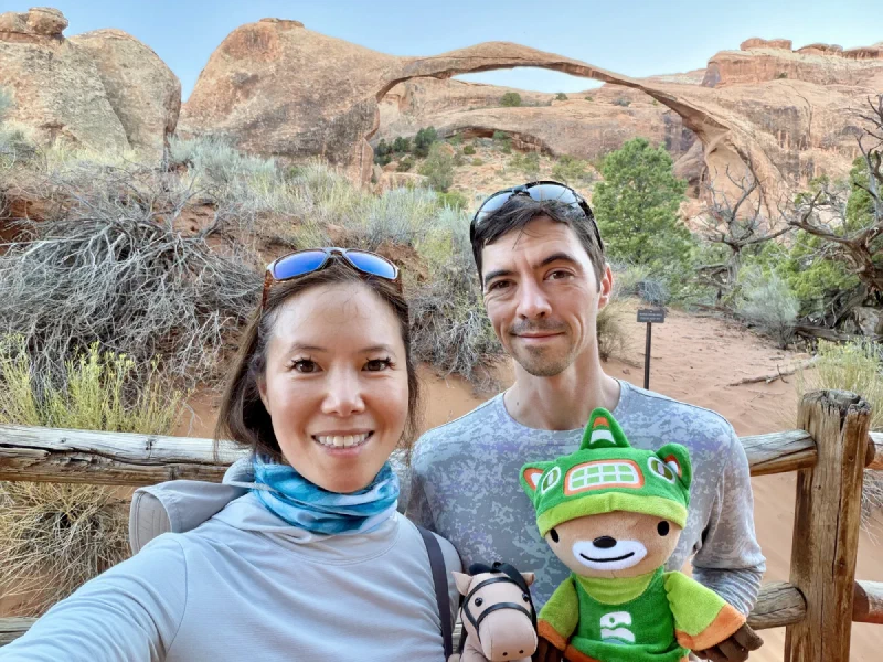 Pokin and Nicholas smiling at the camera with Sumi and Chestnut the toy horse held up in front, Landscape Arch spanning the sky behind