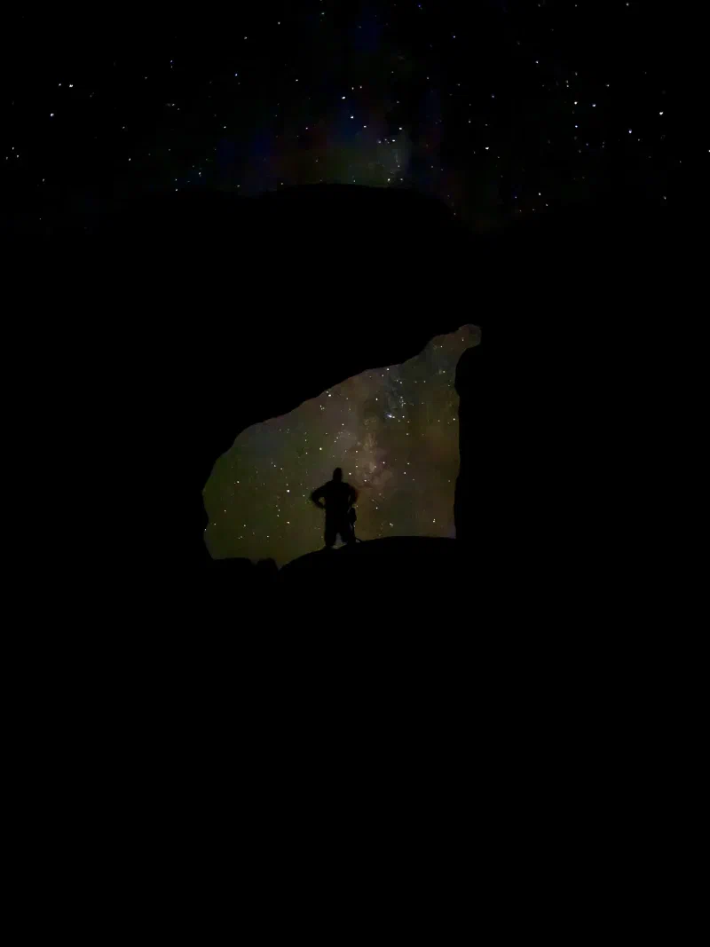 A very dark phone photo of a silhouetted figure standing inside the arch opening, with a murky glow and scattered stars